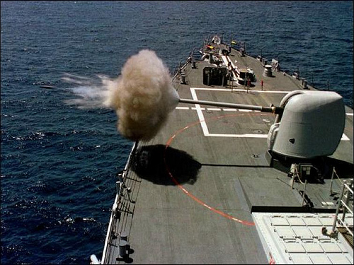 A destroyer firing her forward gun, cigar-shaped muzzle blast extending out over the bow