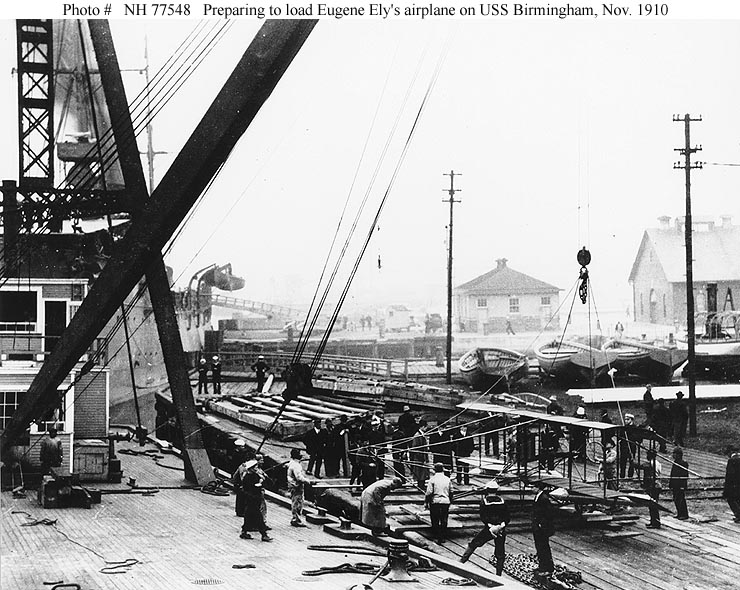 A floating crane lifts Eugene Ely's Curtiss pusher airplane from the Norfolk Navy Yard pier, with USS Birmingham partially visible at left
