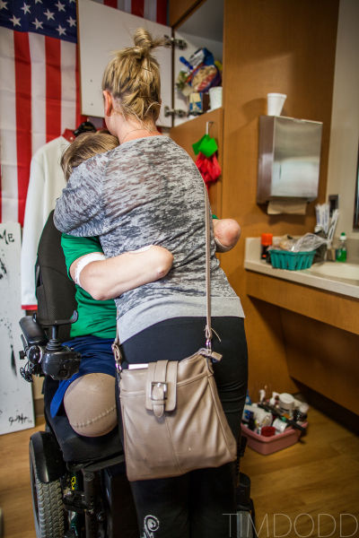 His girlfriend bending down to hold Taylor Morris in his wheelchair, an American flag hanging on the wall behind them