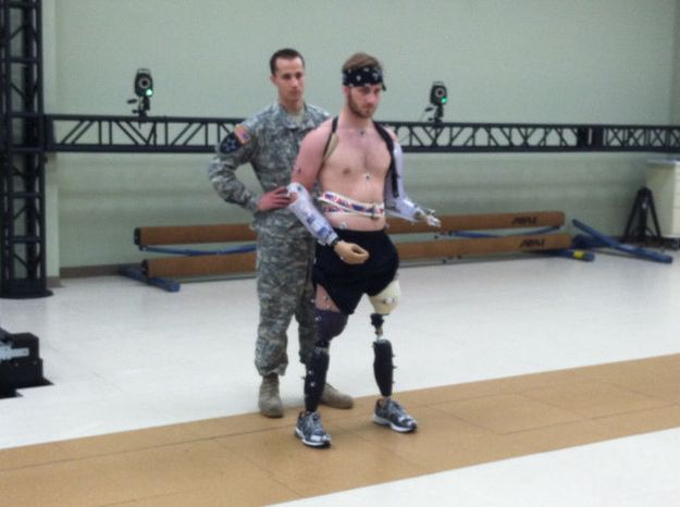 Taylor Morris walking on two prosthetic legs in the rehabilitation gym, a soldier steadying him from behind